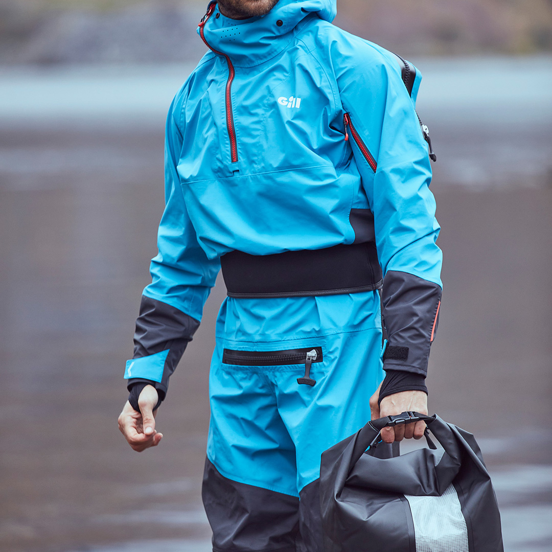 Man wearing a Gill Blue Jay Verso Drysuit and holding a Dry Cylinder Bag next to a lake