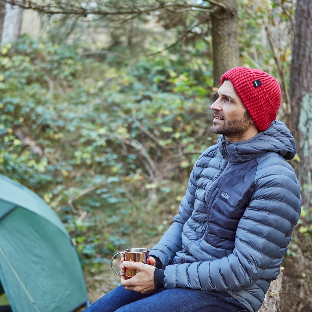 A man sat on a tree trunk drinking coffee wearing a Gill Fitzroy Jacket and red Seafarer beanie