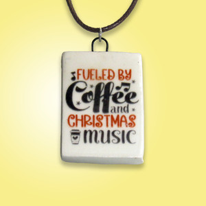 A handmade Ceramic Christmas necklace pendant with the words coffee & Christmas music. On a black cord necklace. Shown on a yellow background. A handmade Ceramic Christmas necklace pendant with the words coffee & Christmas music. On a black cord necklace. Shown on a yellow background.