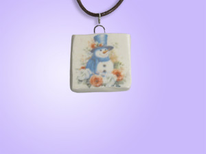 A Handmade Snowman with Blue Top Hat and Scarf Ceramic Pendant Necklace. With black cord. With purple background. A Handmade Snowman with Blue Top Hat and Scarf Ceramic Pendant Necklace. With black cord. With purple background.