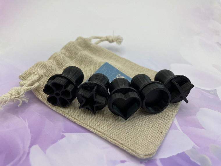 A set of 5 mini clay stamps, including a heart, star, flower, an x & an o. On top of a small burlap pull together bag. On a purple floral background.
