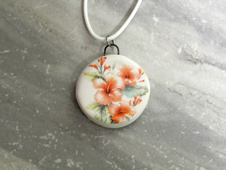 A handmade round peach hibiscus flowers and leaves ceramic pendant necklace. On a white cord necklace. With a gray marble look background.