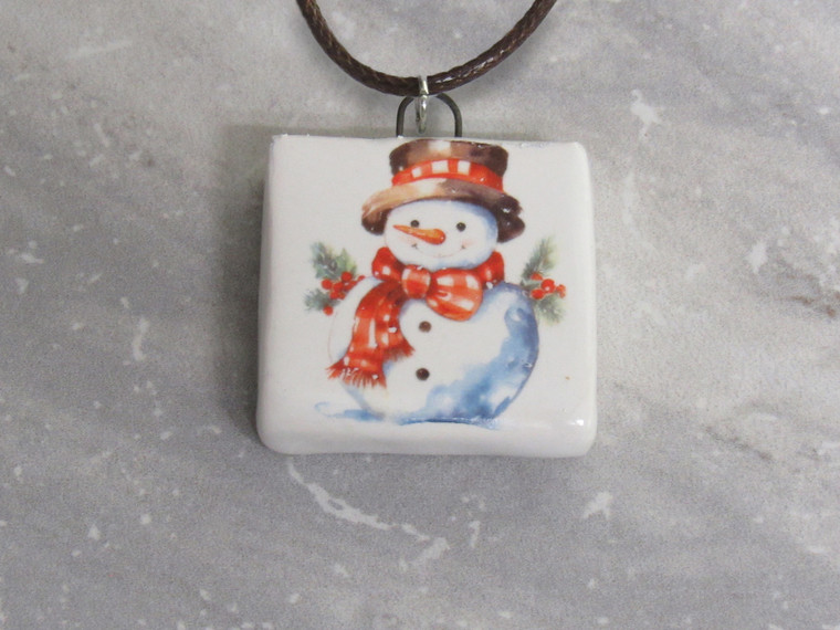 A handmade adorable Snowman with Carrot Nose and Red Scarf square ceramic necklace. On a black cord necklace. With a gray marble look background.
