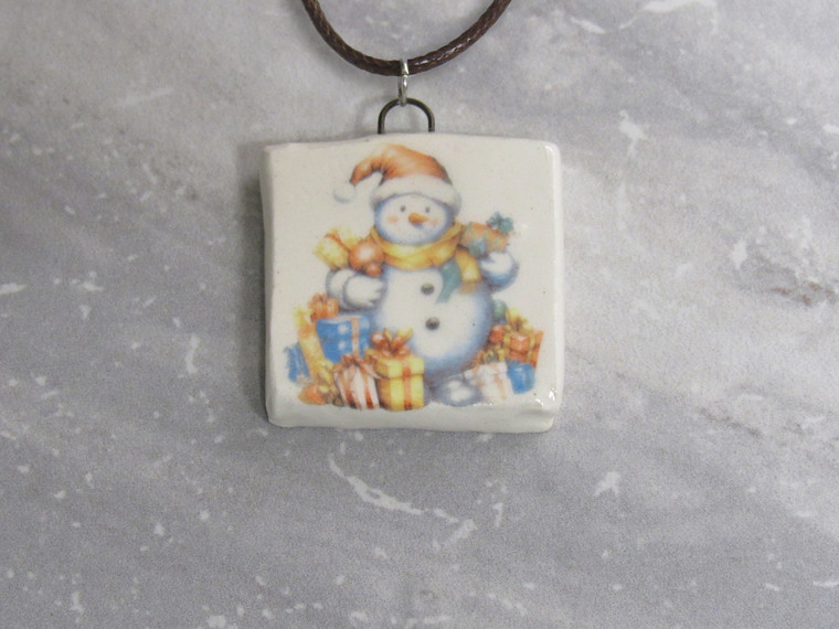 A handmade snowman with packages square ceramic pendant necklace. On a black cord necklace. With a gray marble look background.