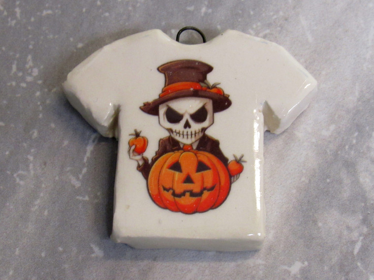 A handmade ceramic Tshirt shaped Halloween necklace. Featuring a skeleton in a Suit holding a Jackolantern. On a gray marble look background.