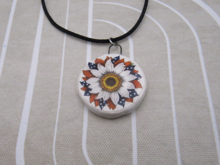 A handmade red and blue Patriotic Daisy Quilt look ceramic pendant necklace. On a black cord necklace.  On a beige and white background.