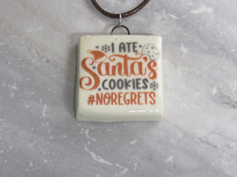 A handmade fun Christmas Ceramic square shaped pendant necklace. With the words "I ate Santa's Cookies no Regrets". On a black cord necklace. With a gray marble look background.
