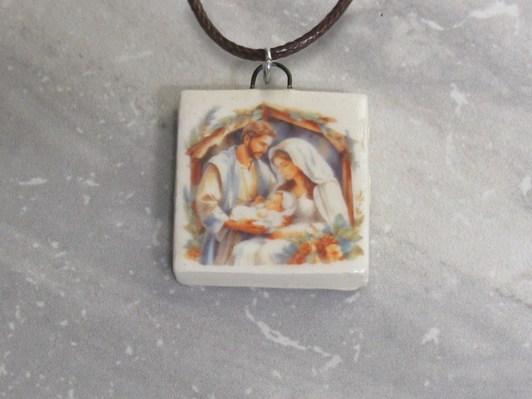 A handmade square ceramic pendant necklace with Mary, Joseph, and Baby Jesus. On a black cord necklace. With a gray marble look background. A handmade square ceramic pendant necklace with Mary, Joseph, and baby Jesus. On a black cord necklace.