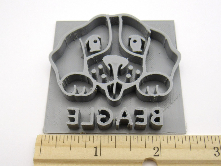 A Beagle mug clay pottery stamp. With a Beagle head with paws on top of the word Beagle. With ruler. Showing approximately 2.5 inches wide.