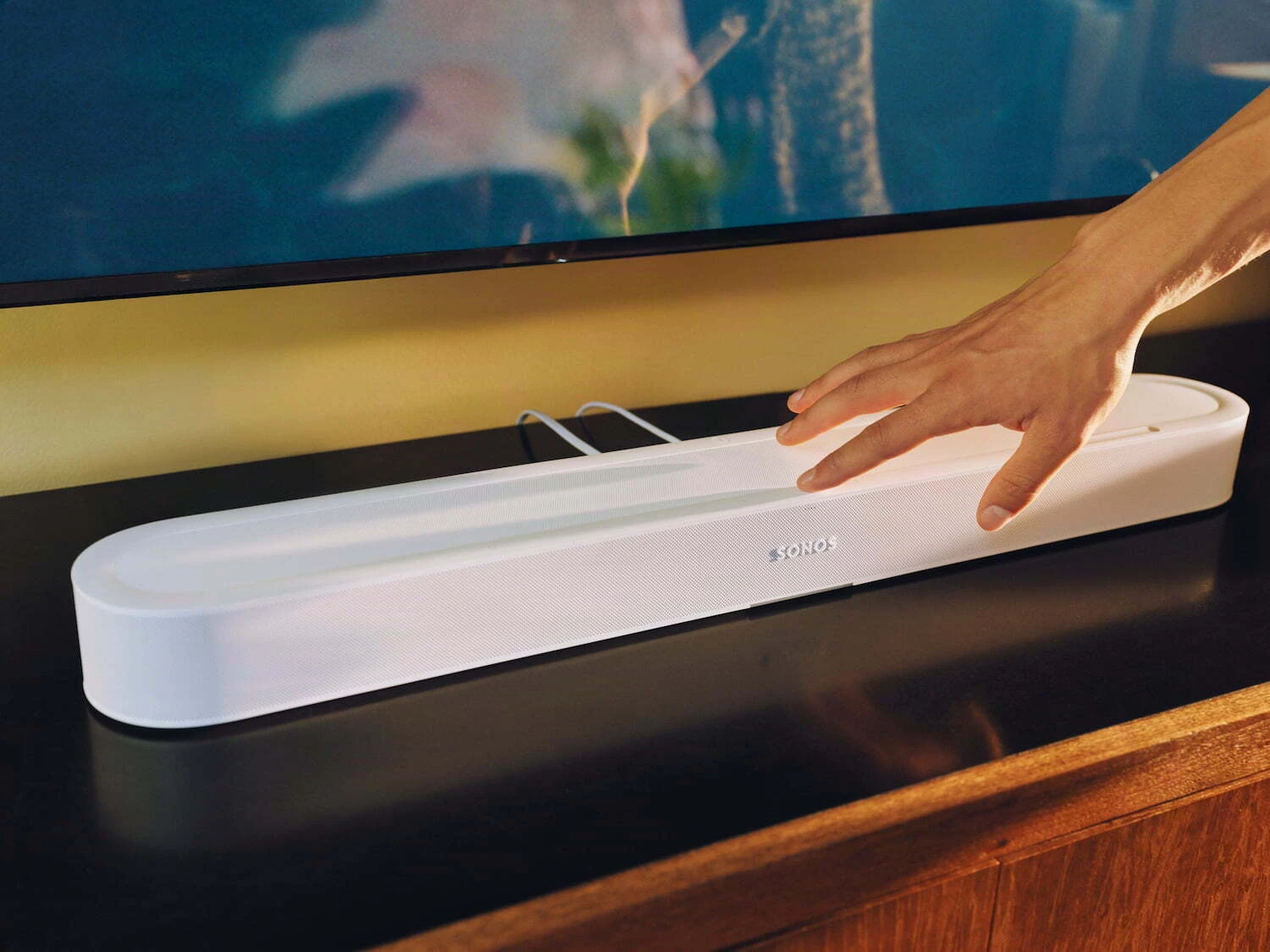 Sonos Beam Ultra soundbar in White on a TV console, hand reaching to use the touch controls
