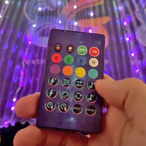 Remote Controlled LED Wings LED Cape with Rainbow Lights, showing a hand holding a remote control with colorful buttons in front of glowing LED wings and cape in a dark setting.