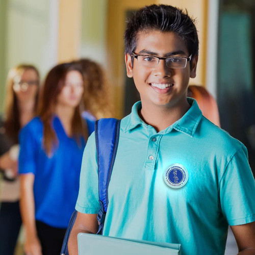 Multicolor Flashing Light Up Pin-on LED 2" Circle Badge, shown on a student wearing a teal shirt and glasses, in a school hallway with other students in the background.