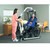 A caregiver using an Invacare Reliant 450 Battery-Powered Patient Lift to assist an individual in a wheelchair, with a bed and dresser in a well-lit room.