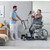 A caregiver assists a patient using a motorized Invacare ISA XPLUS Stand-Up Electric Patient Lift in a bright, spacious room, with a bed and modern wheelchair nearby.