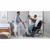 A caregiver assists a patient using an Invacare ISA COMPACT Stand-up Electric Patient Lift next to a wheelchair in a modern, well-lit room.