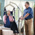 A caregiver assists an elderly person in a sling lift attached to an Invacare 9805P Hydraulic Patient Lift, positioned next to a bed in a brightly lit room.