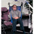 A man is seated in a blue patient transfer sling, designed for mobility assistance, next to a wheeled, cushioned chair in a bright indoor setting.