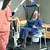 A healthcare worker operates a lift to assist an elderly patient seated on a commode chair in a bedroom setting. A healthcare worker operates a lift to assist an elderly patient seated on a commode chair in a bedroom setting.