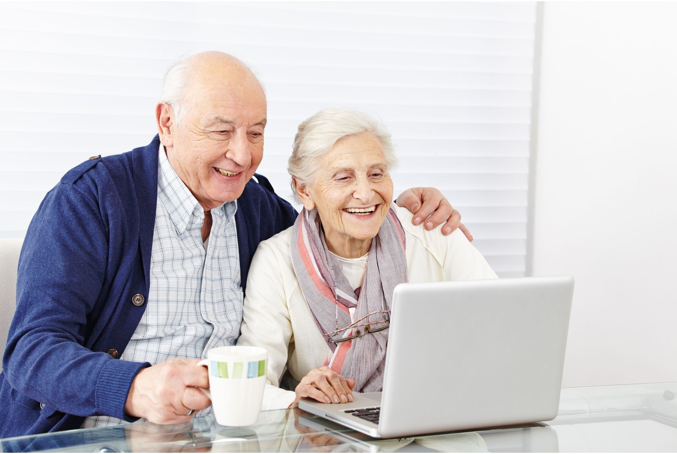 Smiling elderly couple embrace at a table as they focus on a laptop computer screen