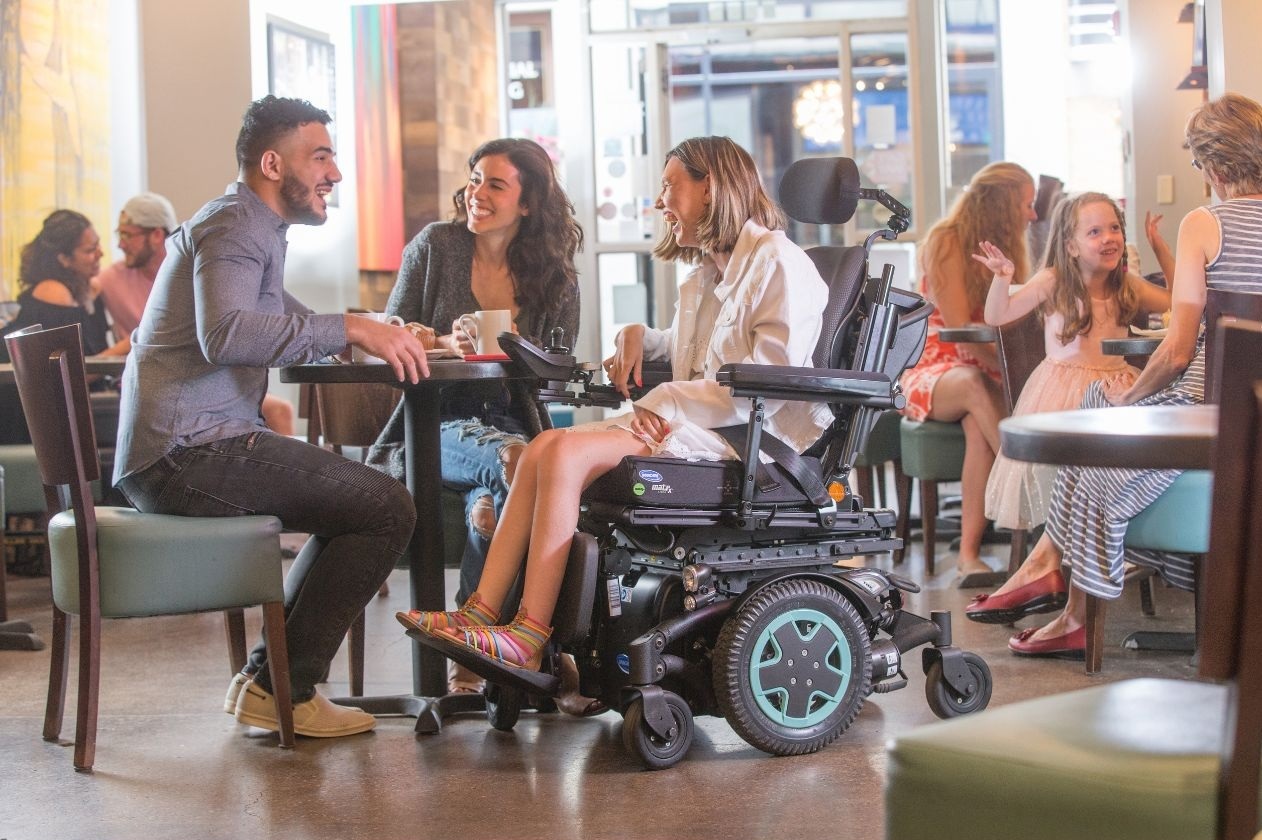 A person seated in a black Invacare TDX SP2 electric wheelchair with green-accented wheels is interacting with others at a modern cafe. The wheelchair features a joystick and padded seat. Several people are gathered around a table, engaged in conversation, with other patrons visible in the background.