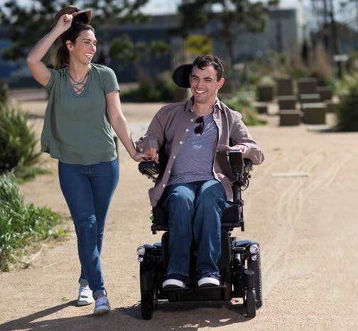 Two people outdoors on a sunny day, one walking and holding hands with the other who is seated in an Invacare TDX SP2 Electric Wheelchair. They are on a paved path surrounded by greenery, with buildings in the background.