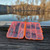 Open orange Saga mini tackle box displaying fishing lures, hooks, and soft plastics on wooden dock by water