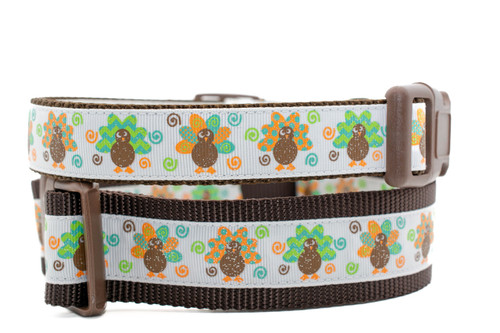 Dog collar with a colorful Thanksgiving theme. The collar features a repetitive pattern of small, funky turkeys with brown bodies and vibrant, sparkly tail feathers in orange, green, and blue swirl designs. The collar has brown edges, a brown plastic buckle, and a silver metal D-ring.
