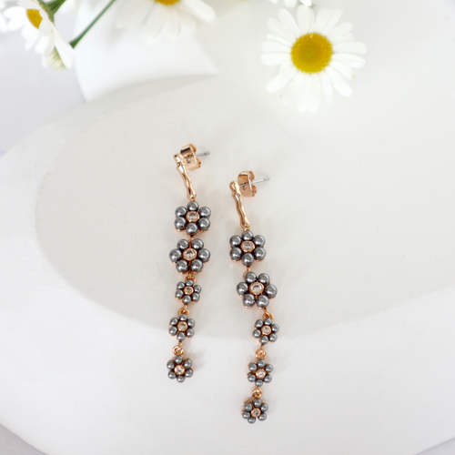 Pair of gold drop earrings with cascading grey pearl daisy designs displayed on a white surface, styled with fresh daisies in the background. Pair of gold drop earrings with cascading grey pearl daisy designs displayed on a white surface, styled with fresh daisies in the background.