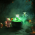  Three plush toys are brewing a glowing green potion in a large cauldron in a spooky, foggy setting with pumpkins and a gate. 
