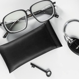 A black PU pouch beside silver glasses, a key, and a keyring. The pouch has a simple design and a logo.