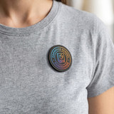 A round, multi-coloured magnetic badge worn on a grey shirt, featuring a logo and text design.