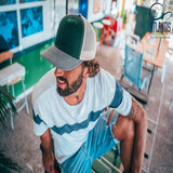 A green and beige trucker cap is worn by a man with curly hair, seated in a casual indoor setting.