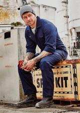 A man wearing a navy blue cotton drill overall and grey beanie, sitting on a weathered crate.