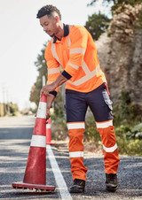 A man in an orange long sleeve polo and navy pants positions a traffic cone on the side of a road.