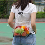 A clear drawstring backpack filled with red apples, green grapes, and a yellow-orange fruit, being worn by a person.