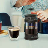 A black coffee plunger with a glass body sits on a table beside a cup of brewed coffee. A black coffee plunger with a glass body sits on a table beside a cup of brewed coffee.