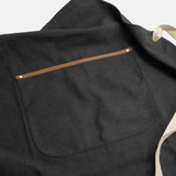 A dark grey canvas apron featuring a large back pocket with a brown leather trim and cream straps. A dark grey canvas apron featuring a large back pocket with a brown leather trim and cream straps.