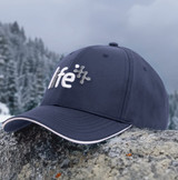 A navy cap with sandwich trim and a logo, resting on a rocky surface with a forested background.