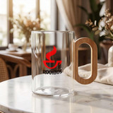 A glass coffee cup with a wooden handle and a red logo, placed on a marble table with a cozy background.