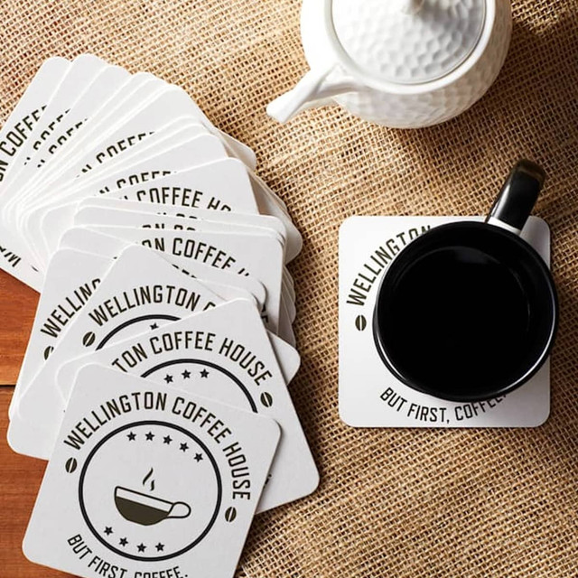 A set of square cardboard coasters in white, featuring a circular design and a coffee-themed logo, next to a black mug.