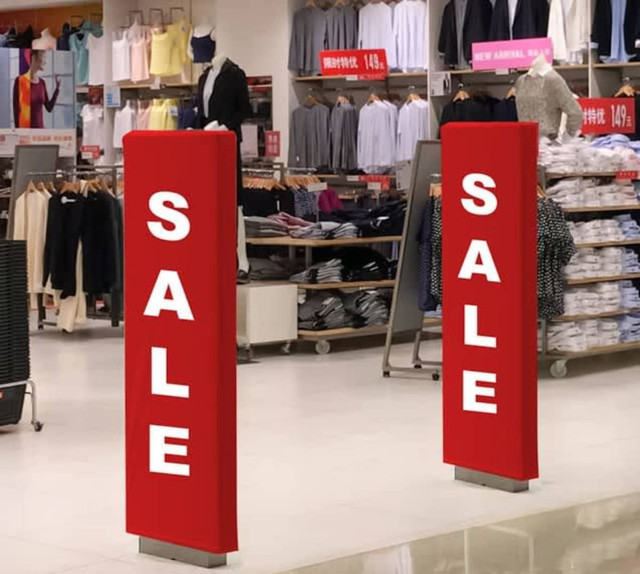Two large red sale signs are positioned in a retail store, surrounded by clothing displays and merchandise.