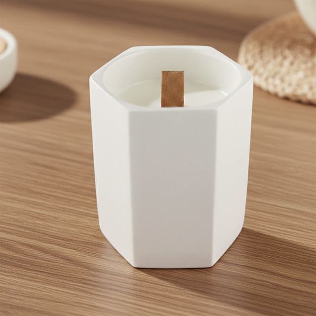 A white hexagon-shaped ceramic candle with a wooden wick on a wooden surface, surrounded by natural elements.