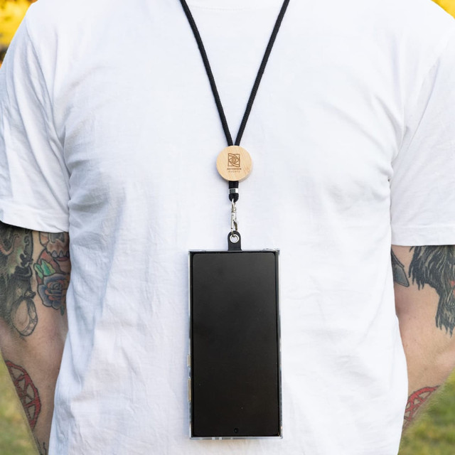 A black neck lanyard with a wooden attachment, holding a smartphone against a plain white shirt. A black neck lanyard with a wooden attachment, holding a smartphone against a plain white shirt.