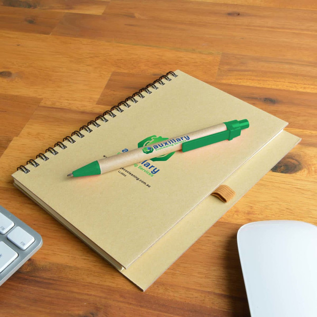 A brown spiral-bound notebook with a green pen placed on a wooden surface next to a keyboard and mouse. A brown spiral-bound notebook with a green pen placed on a wooden surface next to a keyboard and mouse.