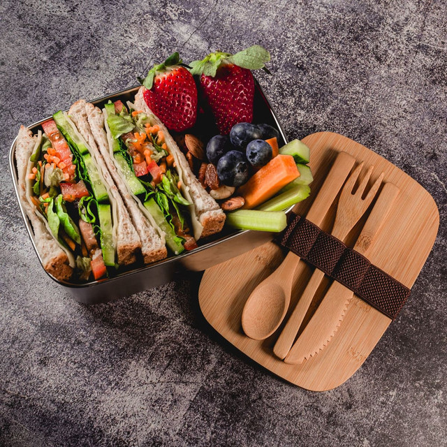 A lunch box featuring wraps with vegetables, strawberries, blueberries, celery, and bamboo utensils. The box has a sleek design.