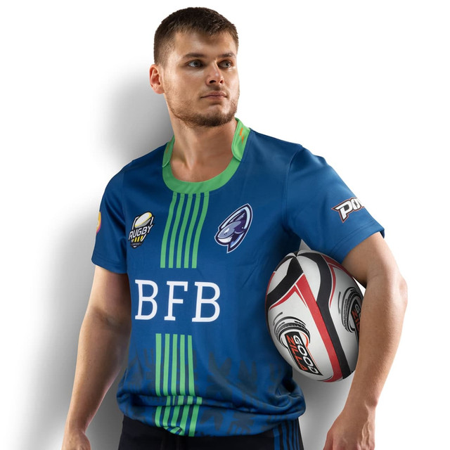 A man in a blue rugby T-shirt with green accents, holding a rugby ball, featuring a logo. A man in a blue rugby T-shirt with green accents, holding a rugby ball, featuring a logo.