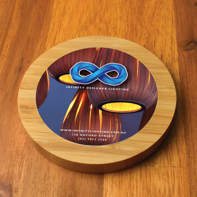A circular bamboo charger featuring a blue logo, set against a wooden table surface. A circular bamboo charger featuring a blue logo, set against a wooden table surface.