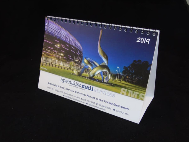 A desk calendar with a printed base featuring an artistic sculpture, displaying months and a logo, predominantly in blue and silver. A desk calendar with a printed base featuring an artistic sculpture, displaying months and a logo, predominantly in blue and silver.