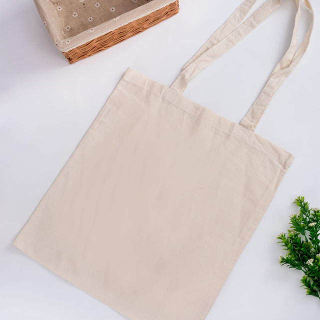 A beige tote bag with long handles, featuring no gusset and a simple design. The image includes a woven basket and greenery.