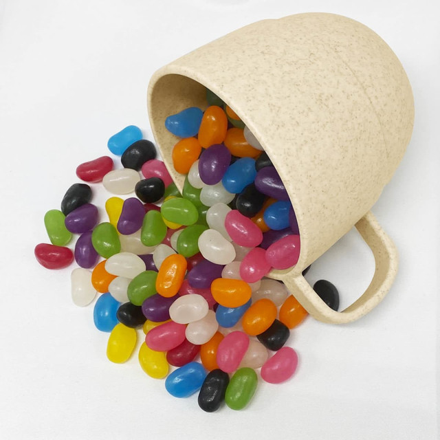 A beige wheat straw cup tipped over, spilling a variety of colourful jelly beans in an assortment of shades.